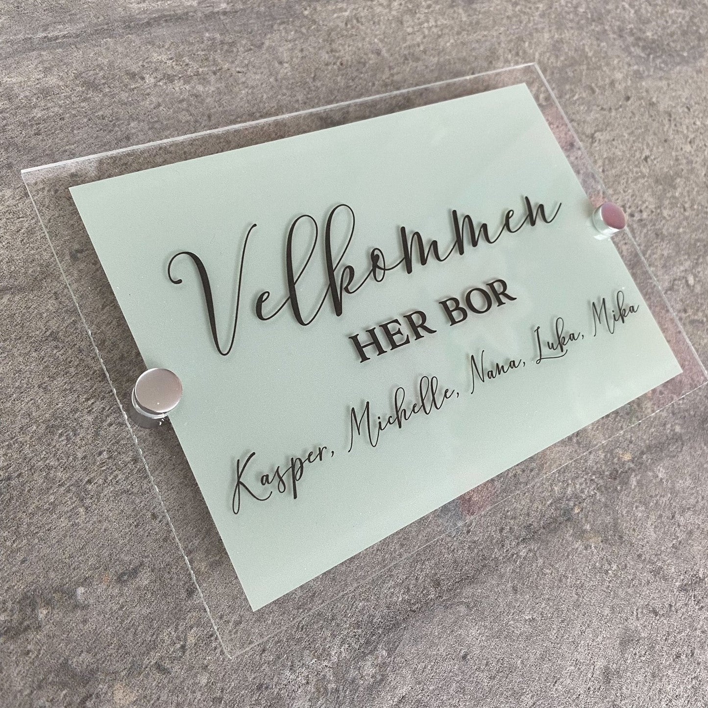 Dette akryl dørskilt er lavet på klar akryl og har en stor firkant i lys grøn på bagsiden. Foran står der velkommen, her bor.